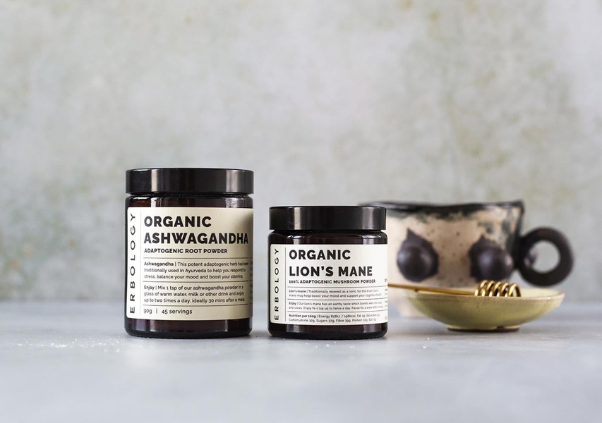 A clean, high-end product photography shot featuring two jars of adaptogenic powders on a neutral surface with a rustic ceramic mug.